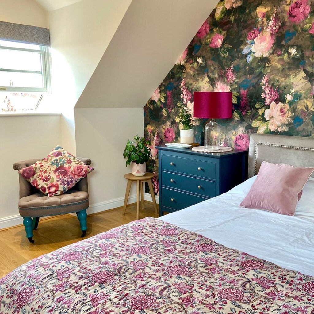 Cheshire interior designer - Colourful bedroom with large bed with pink cushions, blue bedside drawer units, small grey chair and bold floral wallpaper