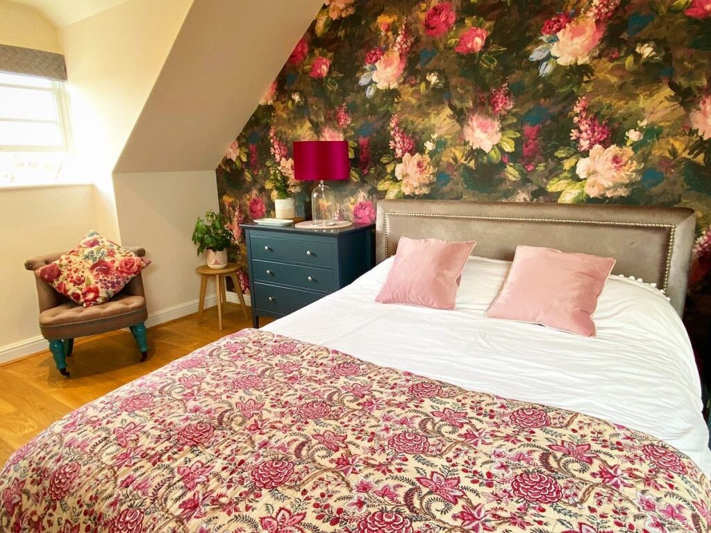 Colourful bedroom with large bed with pink cushions, blue bedside drawer units, small grey chair and bold floral wallpaper - interior design